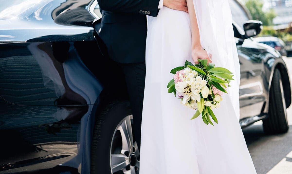 Lady in a white dress holding flowers beside a luxury Bentley wedding chauffeur car in London.